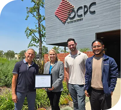 Four people standing outside a building, smiling and holding a framed certificate.